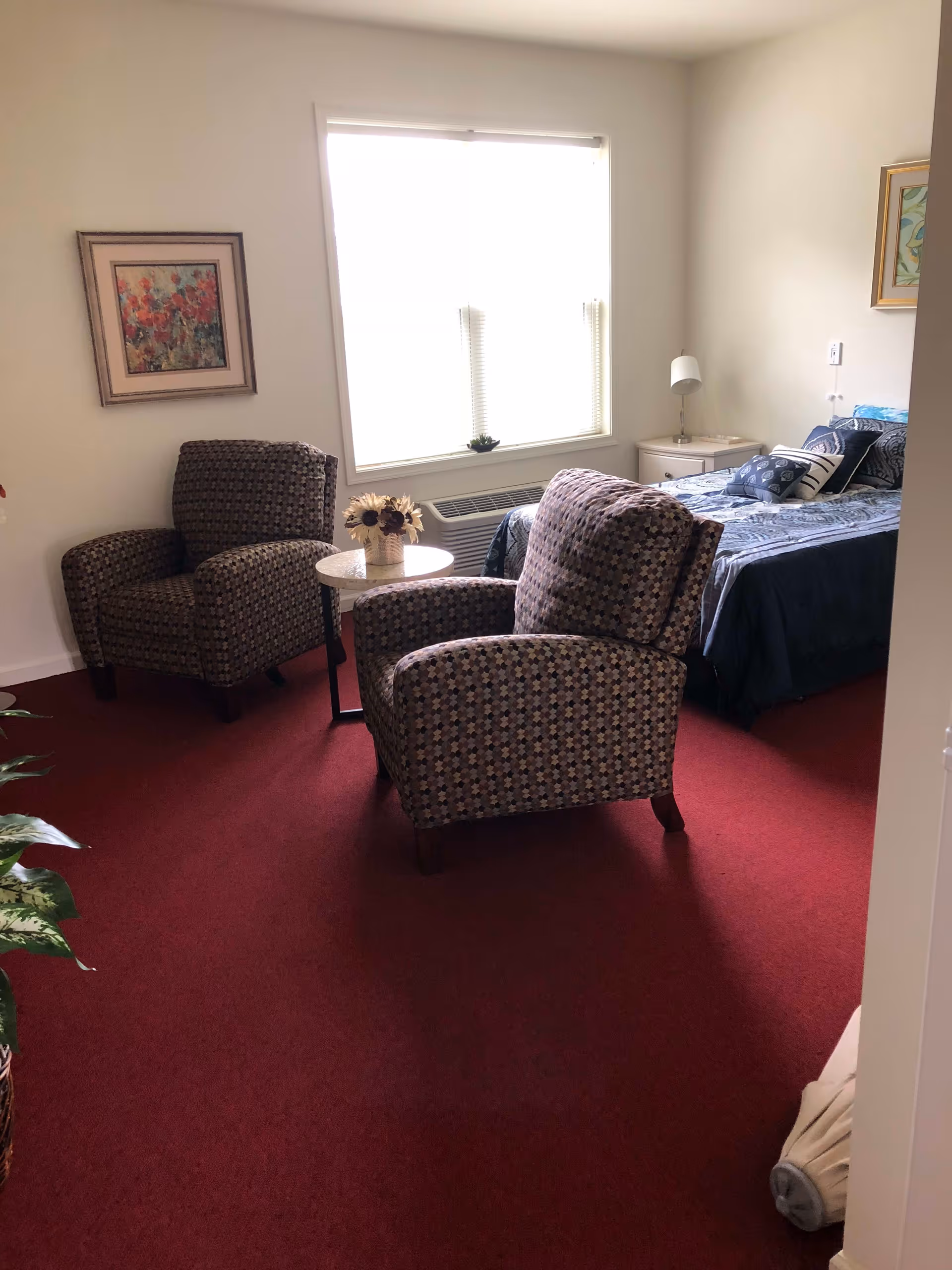A cozy bedroom with two patterned armchairs facing a small round table with a flower arrangement. There is a bed with blue bedding and multiple pillows against the wall, a nightstand with a lamp, a window with blinds, and framed artwork on the walls. The floor is covered with a red carpet.