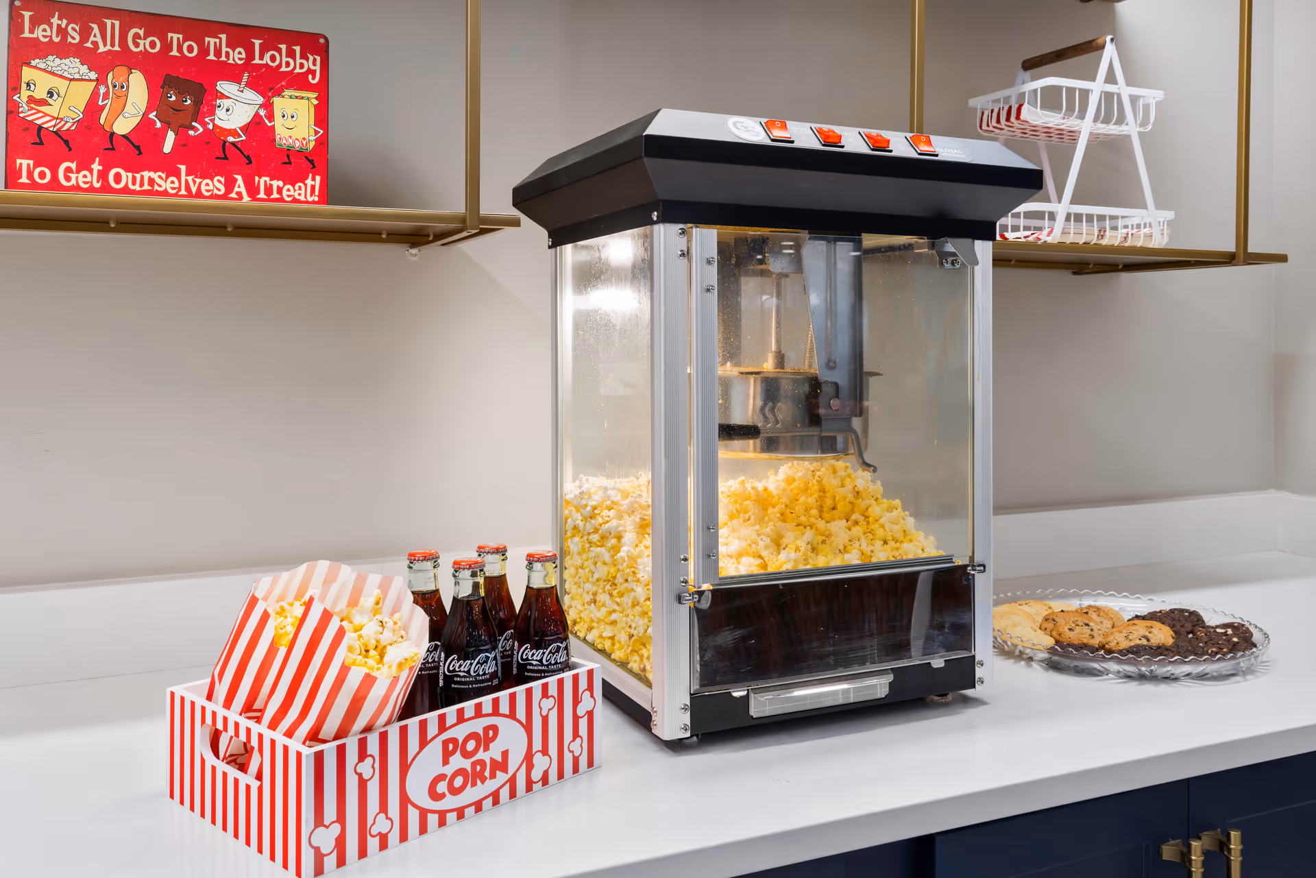 A countertop with a popcorn machine filled with freshly popped popcorn, a striped popcorn container with popcorn, four glass bottles of Coca-Cola in a red and white striped popcorn-themed carrier, and a plate of assorted cookies. A red sign on the wall reads 'Let's All Go To The Lobby To Get Ourselves A Treat!'