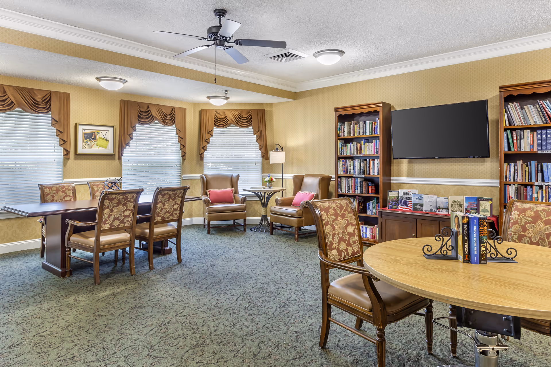 A bright senior living common room with tables, upholstered chairs, bookshelves and a wall-mounted TV.