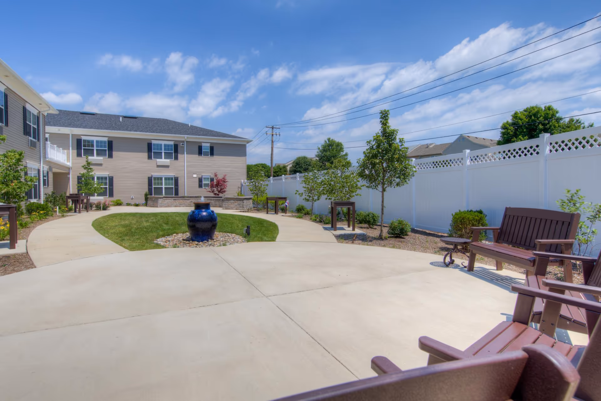 A sunny landscaped courtyard with a central blue urn fountain, curved concrete walkways, benches, small trees and a two-story residential building in the background.