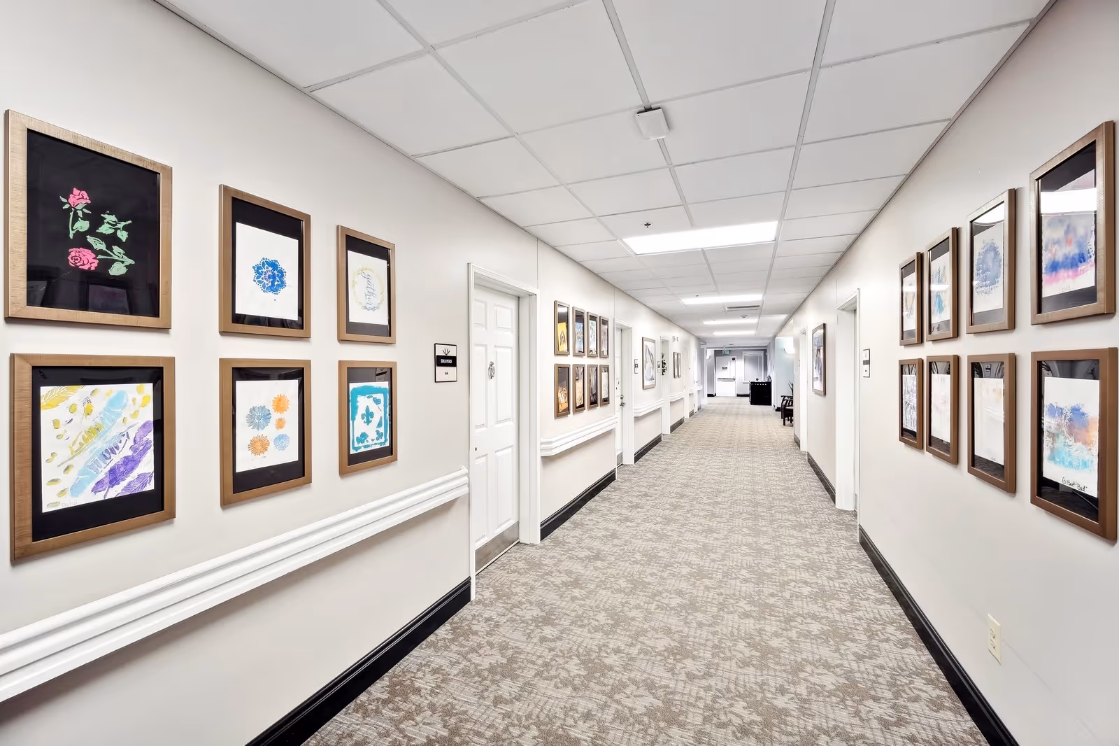 A long, well-lit hallway in a senior living facility with framed artwork hanging on both walls. The walls are painted white, and the floor is covered with a patterned carpet. Several doors line the hallway, and there is a chair and some furniture visible at the far end.