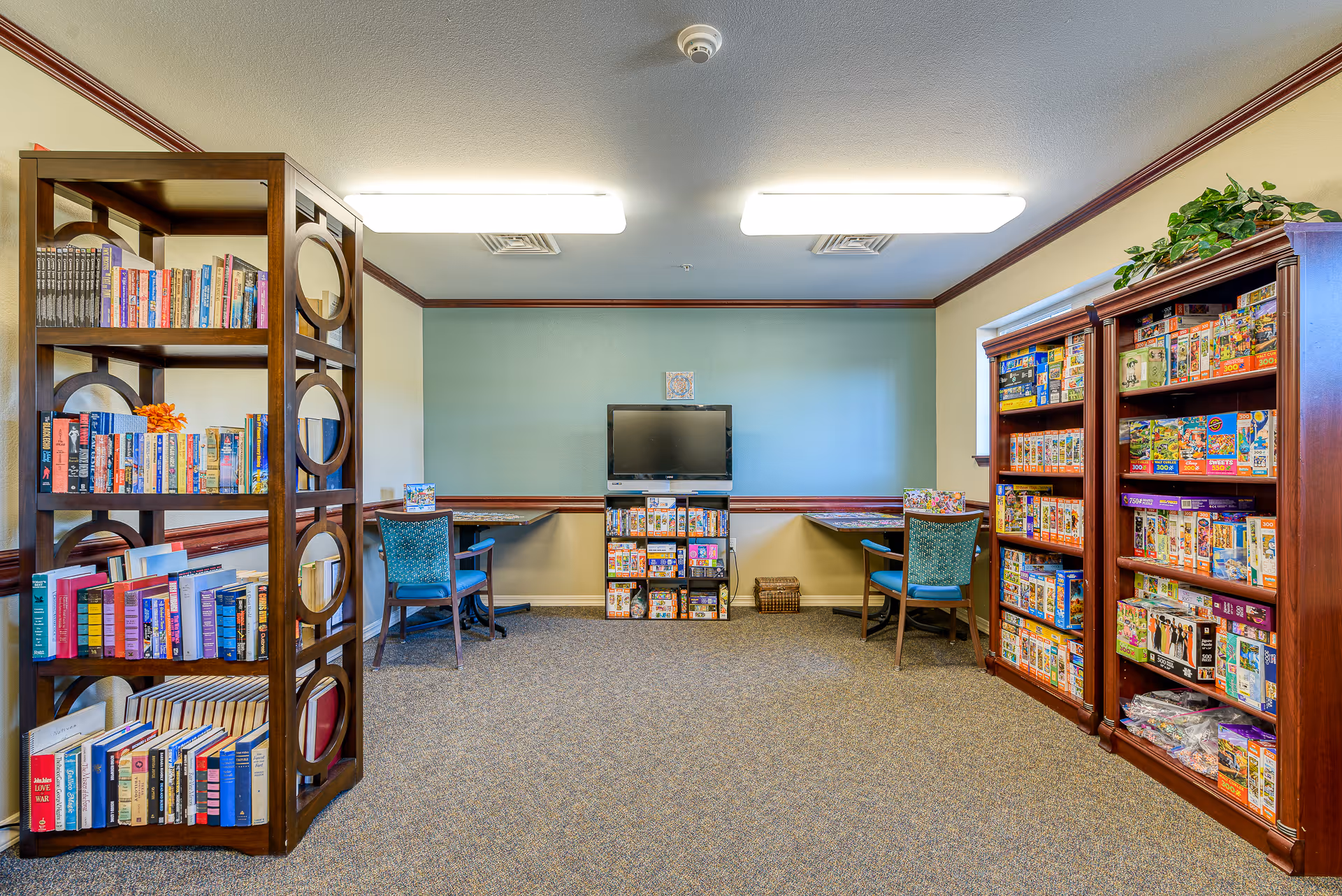 A cozy activity room in an assisted living facility featuring bookshelves filled with books on the left and shelves stocked with puzzles on the right. Two tables with blue chairs are positioned against the back wall, which is painted green and beige. A flat-screen TV is mounted on the wall above a small shelf holding more puzzles. The room is well-lit with ceiling lights and has carpeted flooring.