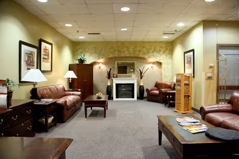 A cozy living room area in a senior living facility with brown leather sofas and armchairs arranged around a central coffee table. The room features a fireplace with decorative vases on either side, framed artwork on the walls, table lamps, and a magazine rack. The walls are painted in warm tones and the ceiling has recessed lighting.