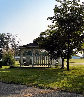 A white wooden gazebo with a picket fence surrounding it, situated on a grassy area with trees nearby and a clear sky in the background.