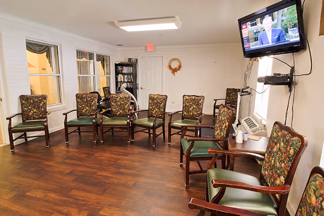 A well-lit room with wooden flooring and a row of chairs with floral patterned backs and green seats arranged along the walls. A flat-screen TV is mounted on the wall to the right, with a small table and an air conditioning unit beneath it. There are windows on the left side and a bookshelf in the back corner. A small decorative wreath hangs on the back wall.