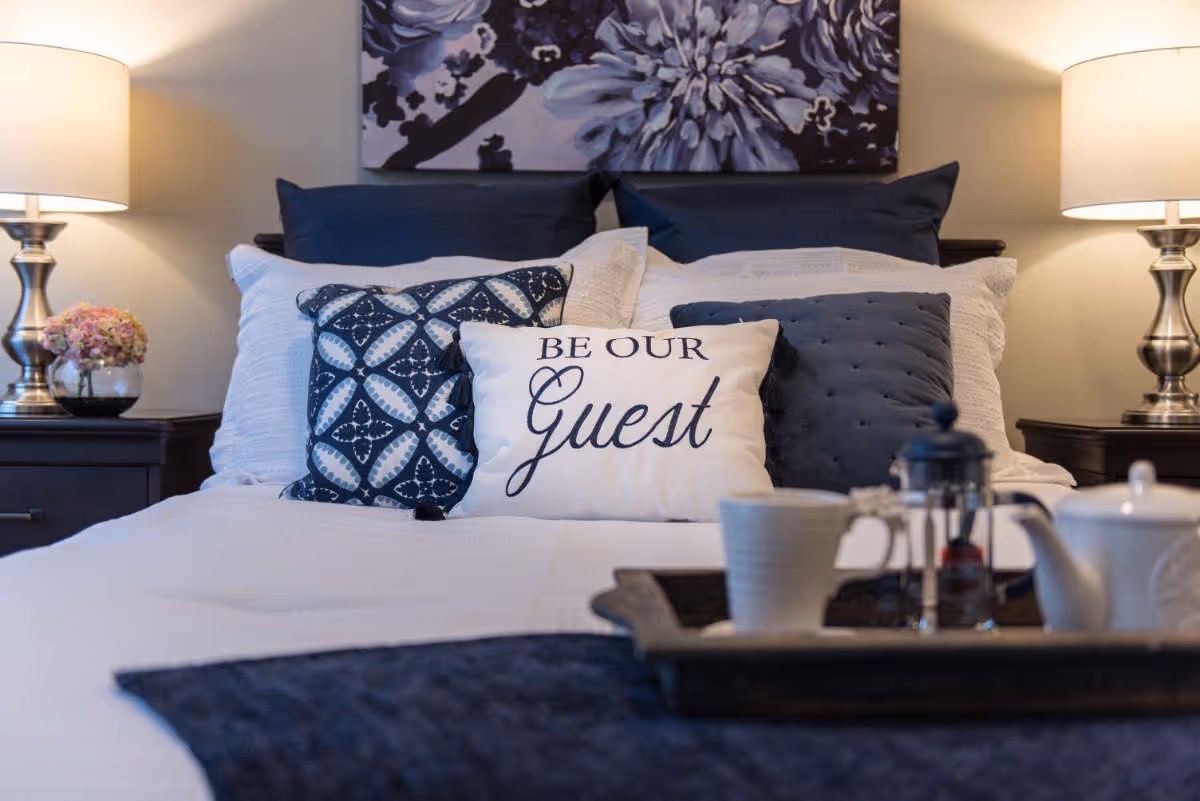 A neatly made bed with multiple pillows including one with the text 'BE OUR Guest'. The bed is flanked by two bedside tables each with a lamp and a small flower arrangement on the left table. A tray with a teapot, cup, and French press is placed on the bed. A large floral artwork hangs on the wall above the bed.