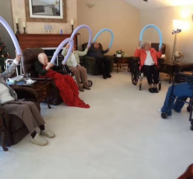 Elderly residents seated in a living room holding curved foam noodles while doing a group exercise.