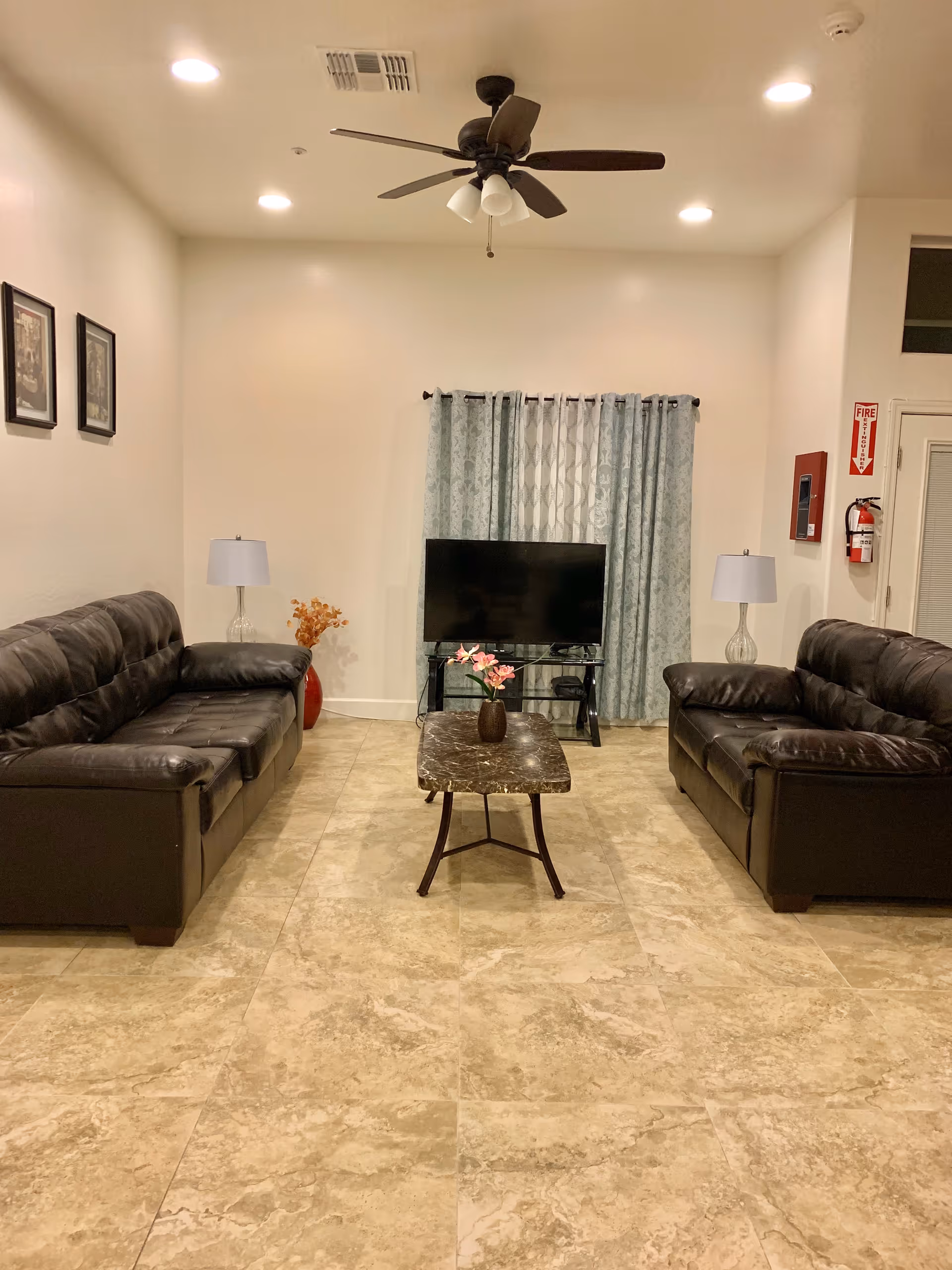 A living room with two black leather sofas facing each other, a small rectangular coffee table with a vase of flowers in the center, a flat-screen TV on a stand against the wall with blue patterned curtains behind it, two table lamps on either side of the sofas, beige tiled floor, ceiling fan with lights, and a fire extinguisher mounted on the wall near a door.