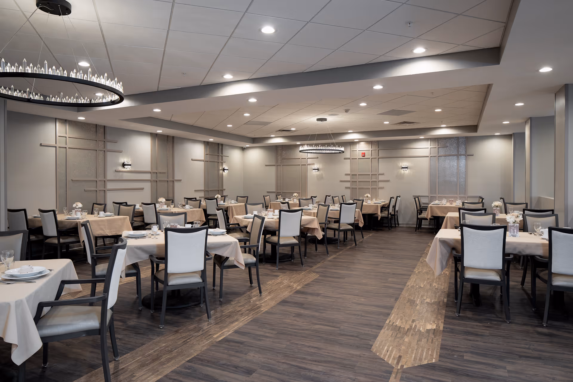 Spacious, neutral-toned dining room with multiple tables set for service and rows of chairs under modern chandeliers.