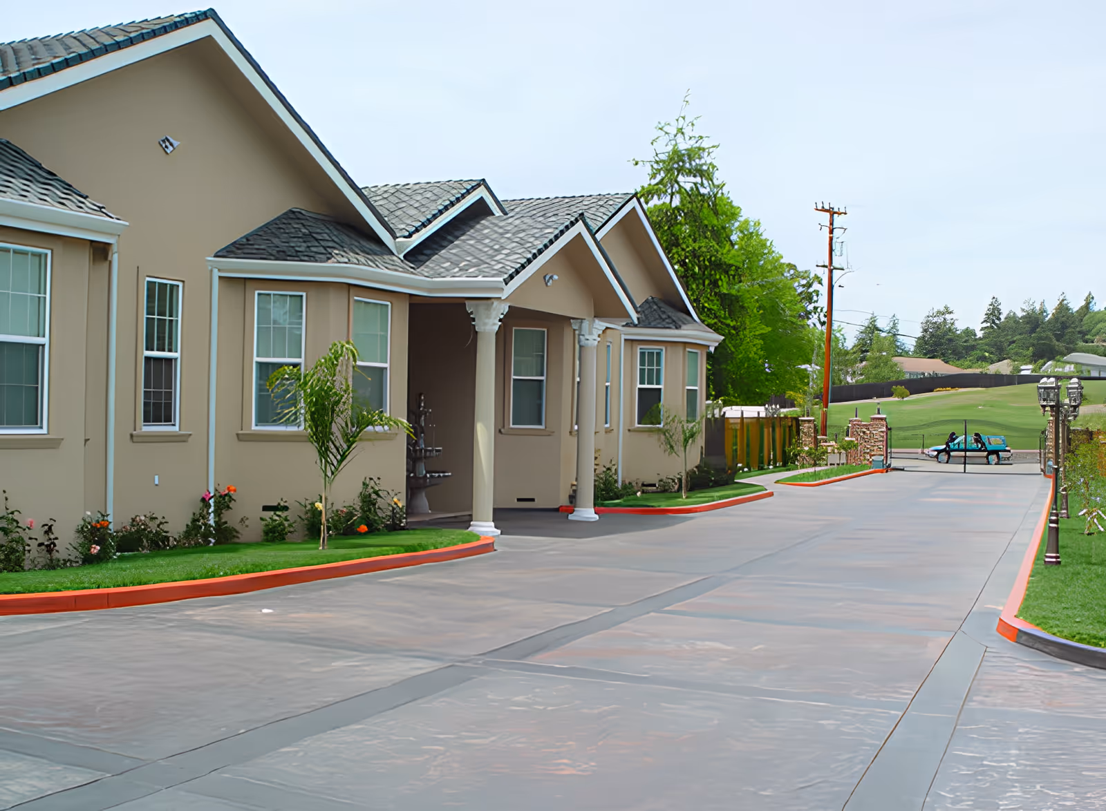 Front exterior of a beige single-story senior living building with a driveway, landscaped lawn, and a small cart in the distance.