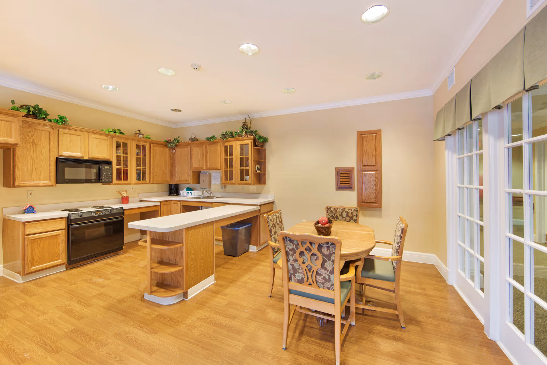 Bright communal kitchen and dining area with oak cabinets, a center island, and a round table with chairs near glass doors.
