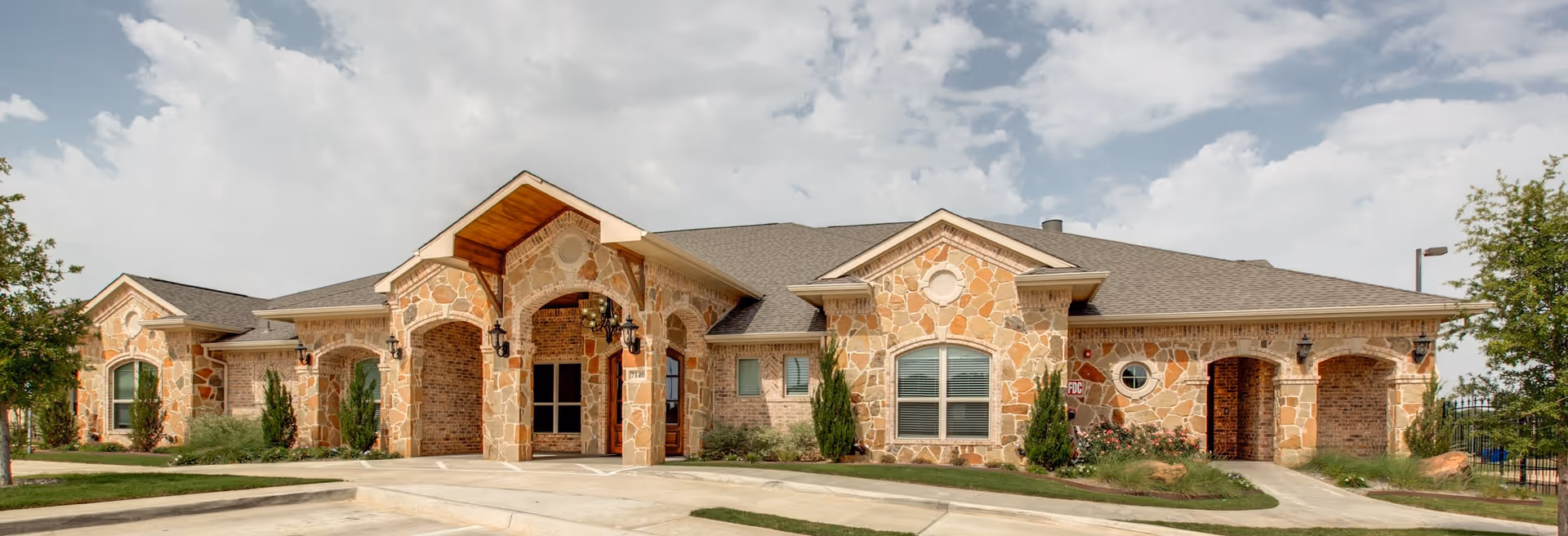 Exterior front view of a single-story stone building with multiple arched entrances and windows, surrounded by landscaped greenery and a paved driveway under a partly cloudy sky.