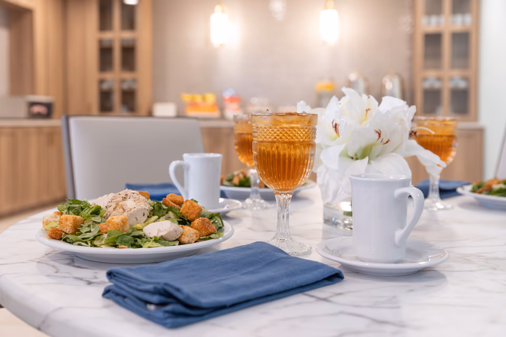A close-up view of a dining table set with plates of salad topped with grilled chicken and croutons, crystal glasses filled with iced tea, white coffee cups on saucers, blue cloth napkins, and a vase with white flowers in the center. The background shows a softly lit kitchen or dining area with wooden cabinets and glass doors.