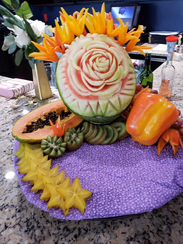 A decorative fruit arrangement on a kitchen counter featuring a watermelon intricately carved with a rose design, surrounded by slices of starfruit, kiwi, papaya, and a whole orange bell pepper. The arrangement is placed on a purple cloth with a floral pattern. In the background, there are bottles, a vase with white flowers, and a computer screen.