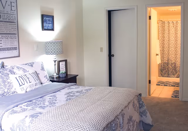 A cozy bedroom with a bed featuring patterned bedding and pillows, a nightstand with a lamp and framed pictures, and a door leading to a bathroom with a shower curtain and bath mats.