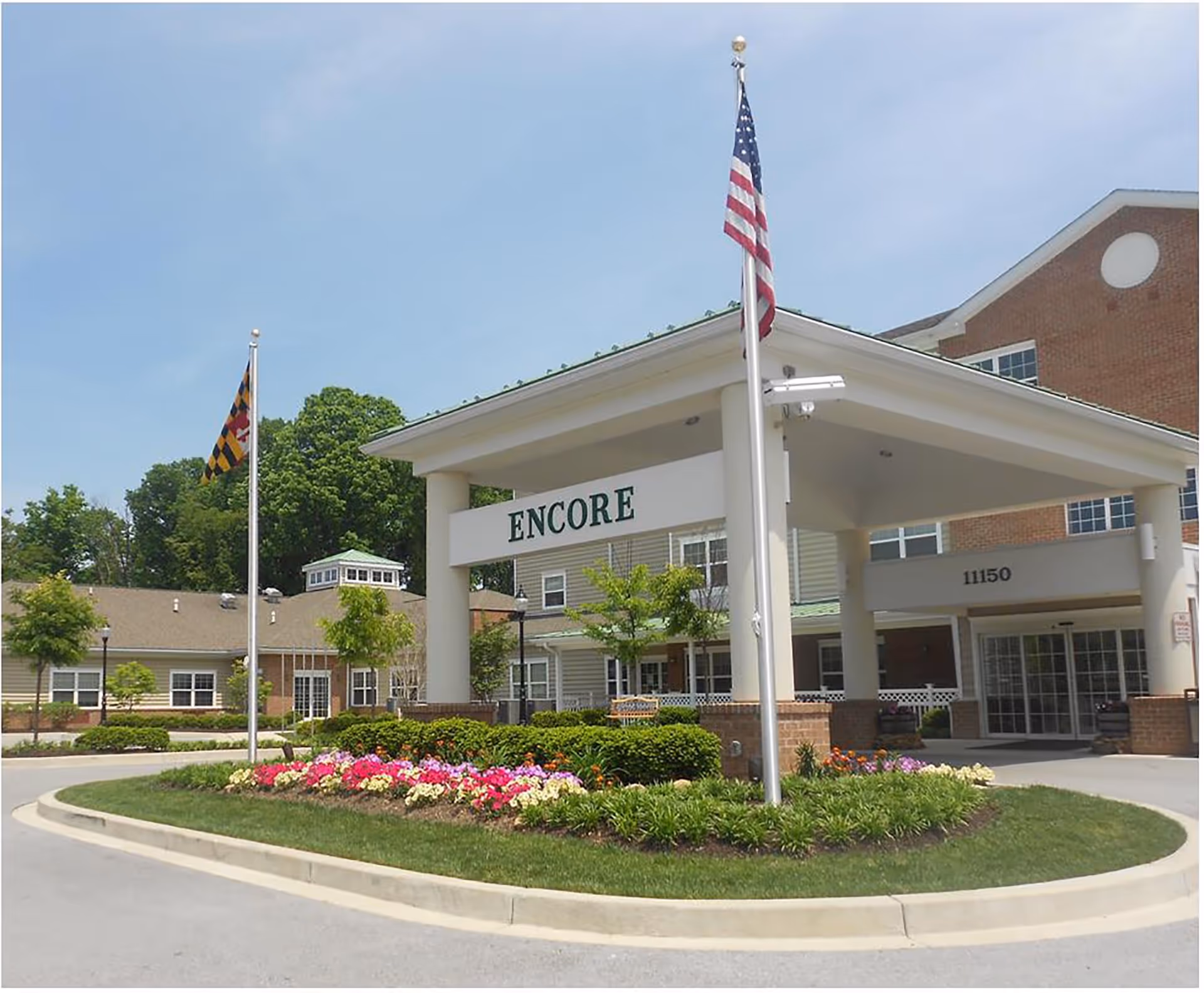 Front entrance of the Lorien Encore senior living facility with a covered driveway, two flagpoles displaying the American and Maryland state flags, and a landscaped flower bed in front.