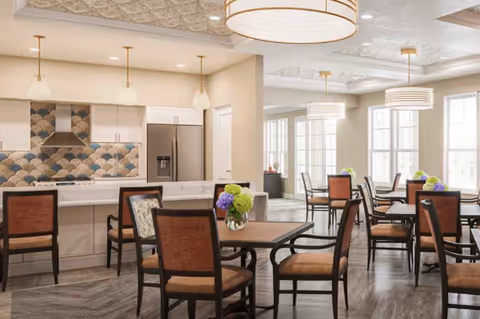 A bright dining area with multiple wooden tables and chairs, each table decorated with a small vase of flowers. The room features large windows allowing natural light to fill the space. In the background, there is a modern kitchen area with a refrigerator, stove, and a decorative tiled backsplash. Pendant lights hang from the ceiling, and the floor has a wood-like finish.