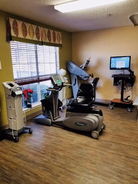 A small exercise room with a recumbent stationary bike, a piece of exercise equipment against the wall, a computer on a mobile stand, and a window with blinds and a decorative valance. The floor is wood-patterned, and the walls are painted beige and green.