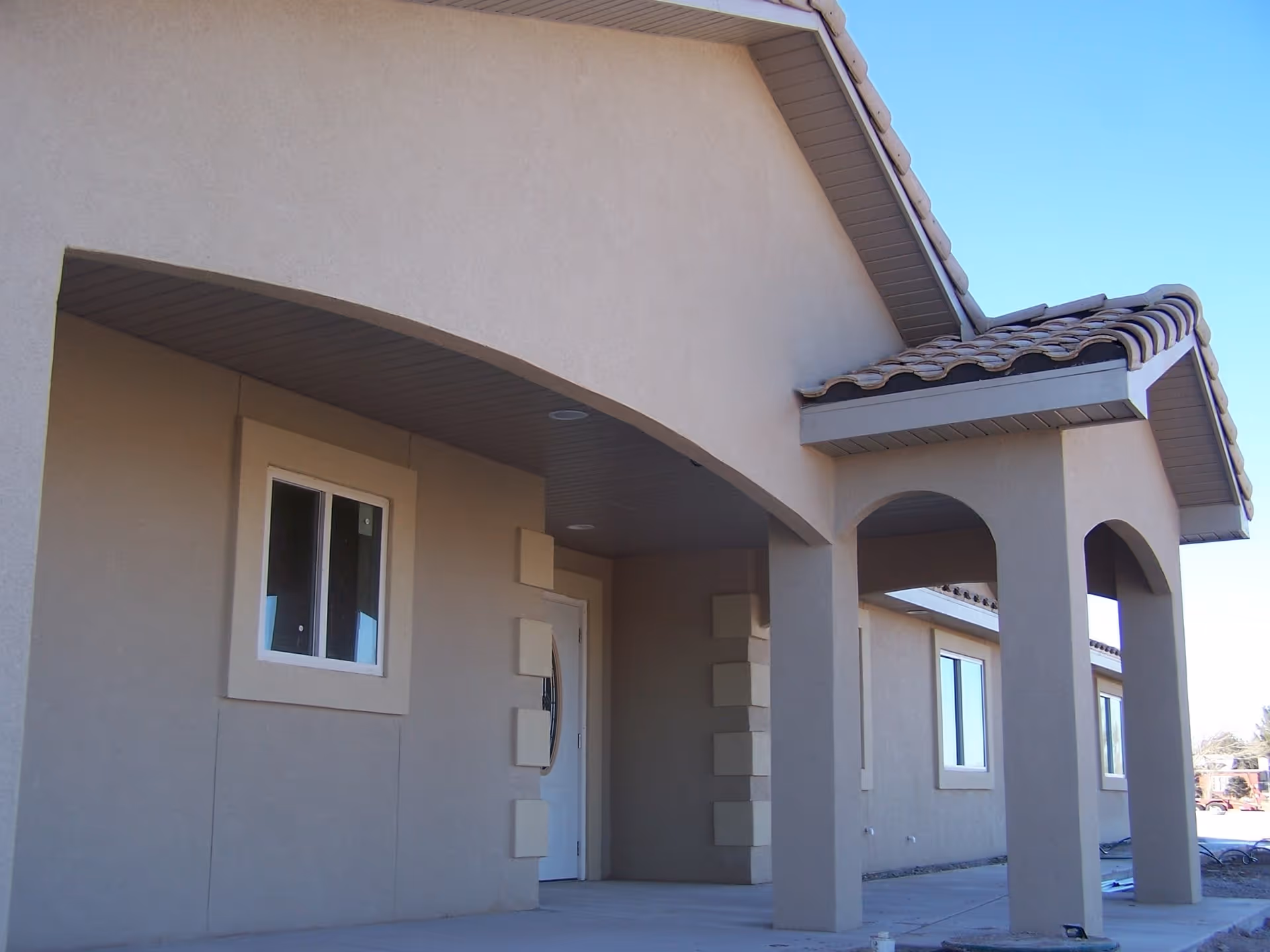 Exterior view of a building with beige stucco walls, a tiled roof, and an arched covered entrance supported by columns under a clear blue sky.