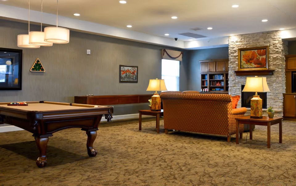A cozy senior living facility common room featuring a pool table, a shuffleboard table along the wall, a patterned sofa with side tables and lamps, a stone fireplace with a floral painting above it, and built-in wooden bookshelves.