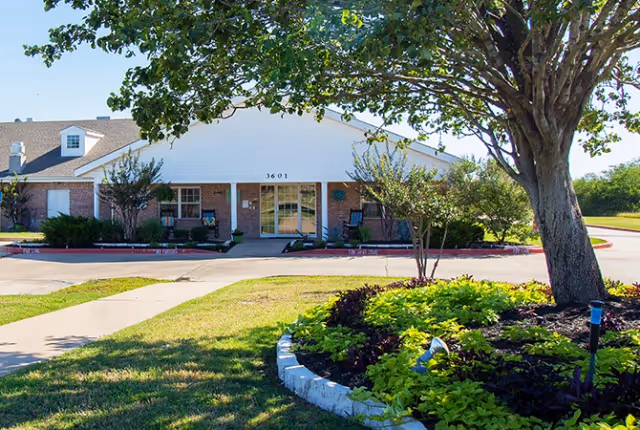 Front exterior of a single-story brick-and-siding senior living building with a central entrance, driveway, and landscaped lawn with trees.