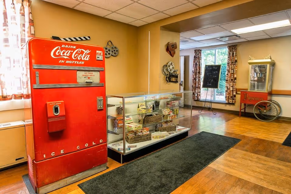 Interior room with a vintage red Coca-Cola vending machine, a glass display case filled with candy and snacks, a popcorn machine on wheels, and a chalkboard sign near a window with patterned curtains. The room has wooden floors and beige walls with movie-themed decorations.