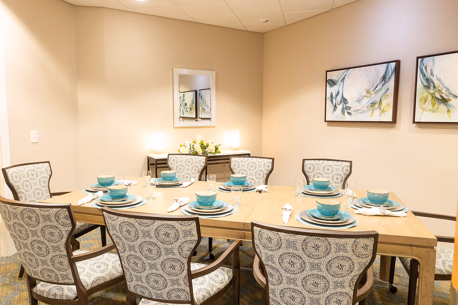 A well-lit dining room with a wooden table set for six with teal dishware and patterned chairs.
