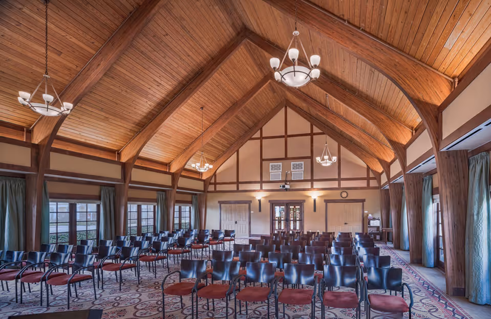 A large room with a high vaulted wooden ceiling and exposed beams, set up with rows of black chairs with red cushions facing a wall with double doors and a projector. The room has large windows with green curtains and patterned carpet flooring.