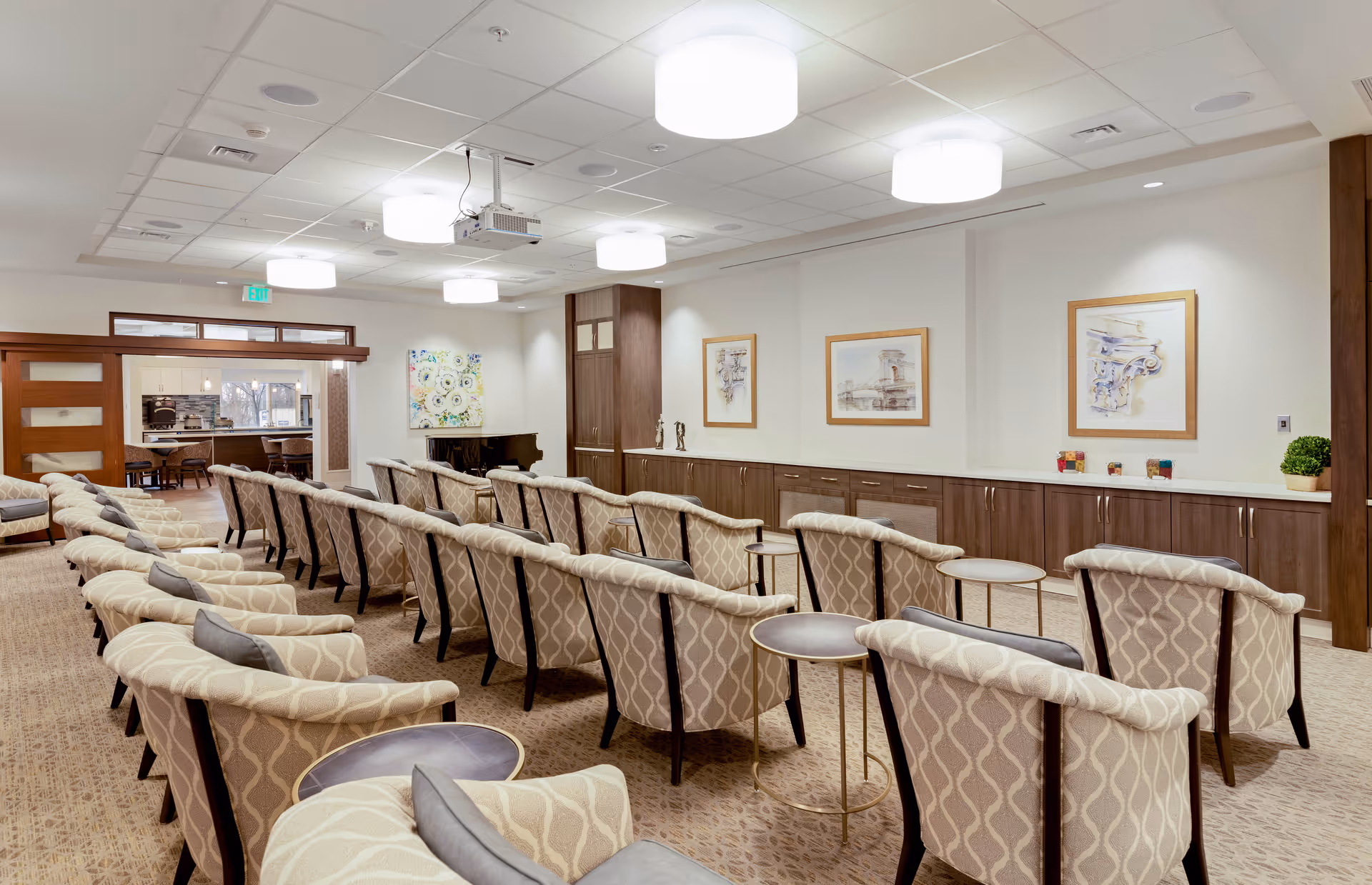 A well-lit meeting or presentation room with rows of patterned armchairs facing a wall with framed artwork. The room has a carpeted floor, modern ceiling lights, and a projector mounted on the ceiling. There are wooden cabinets along the wall and a piano in the corner. An open doorway leads to another room with tables and chairs.