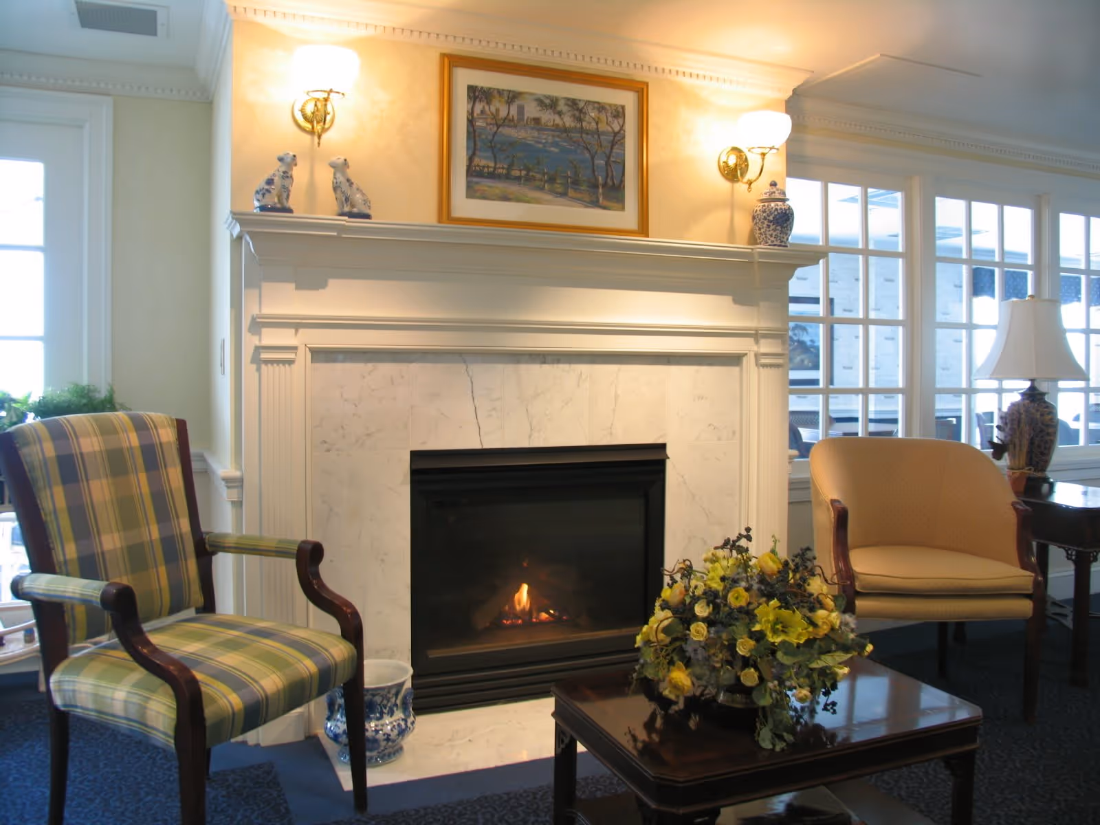 A cozy living room area featuring a lit fireplace with a white marble surround and mantel. On the mantel are two decorative dog figurines, a framed painting, and a blue and white vase. In front of the fireplace is a wooden coffee table with a floral arrangement. To the left is a plaid upholstered armchair, and to the right is a beige upholstered armchair next to a side table with a lamp. Large windows with white frames are in the background, allowing natural light into the room.