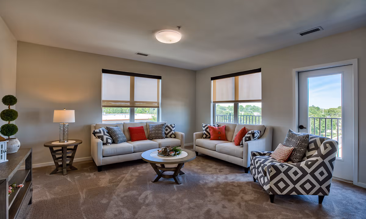 A bright and cozy living room with two beige sofas adorned with patterned and red throw pillows, a patterned armchair, a round wooden coffee table with decorative items, a side table with a lamp, and large windows with beige roller shades letting in natural light. There is a door with a glass panel leading outside.