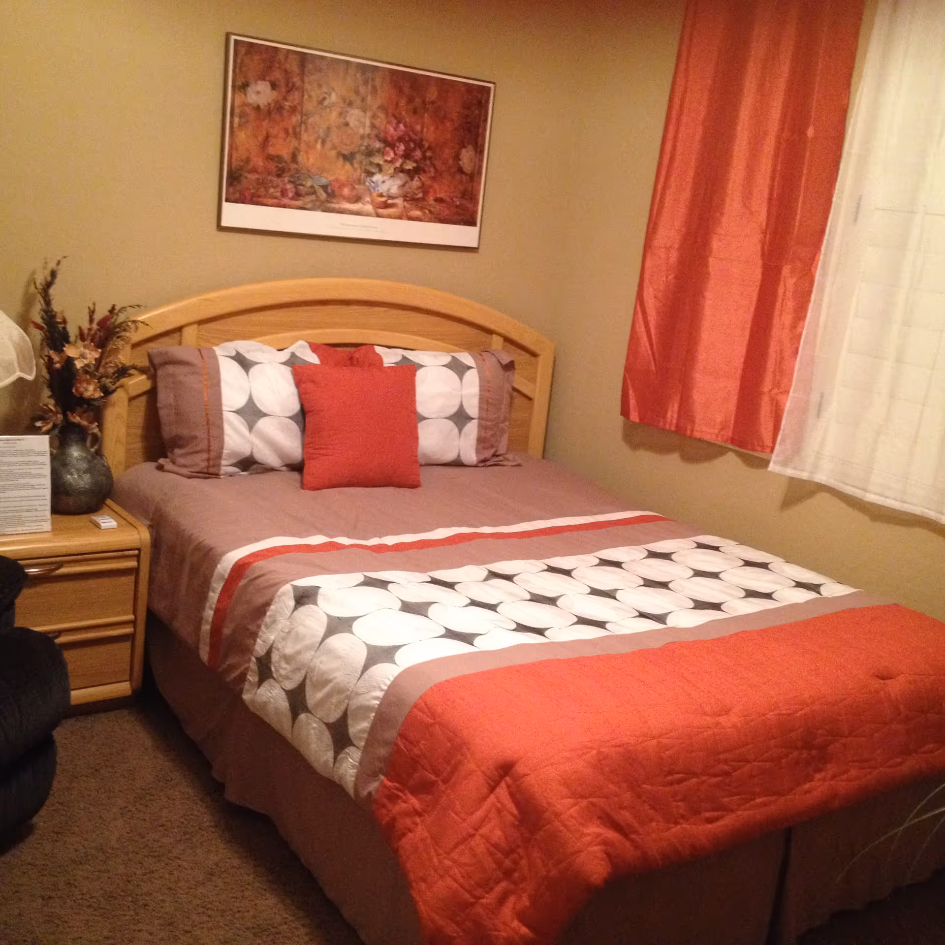 A cozy bedroom with a wooden bed frame and a bed made with a patterned comforter in shades of orange, white, and gray. There are three pillows on the bed, including a solid orange one in the center. A wooden nightstand with a vase of dried flowers and a small lamp is beside the bed. The walls are painted beige, and a framed floral painting hangs above the bed. Orange and white curtains cover the window on the right side.