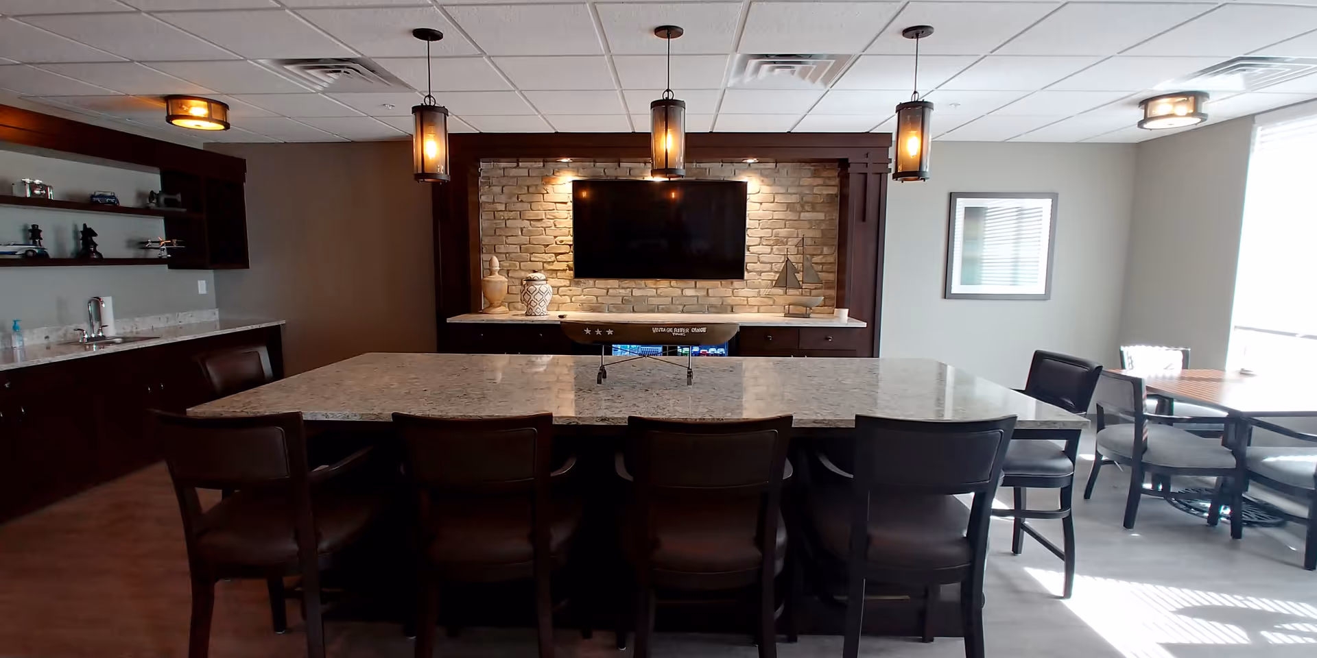 A modern communal dining and lounge area with a large rectangular marble table surrounded by dark wooden chairs. The back wall features a mounted flat-screen TV on a brick accent wall with decorative vases and a small sailboat model on a marble countertop below. Pendant lights hang from the ceiling, and there is a smaller table with chairs near a window letting in natural light.