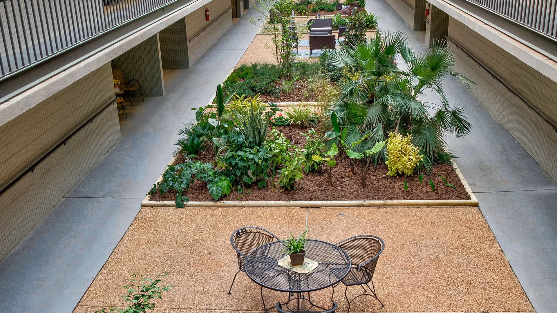 Indoor courtyard area with a central garden bed filled with various green plants and small palm trees. Surrounding the garden bed is a paved walkway with two black metal chairs and a round table with a small potted plant on it. The courtyard is flanked by building walls with balconies and walkways on both sides.