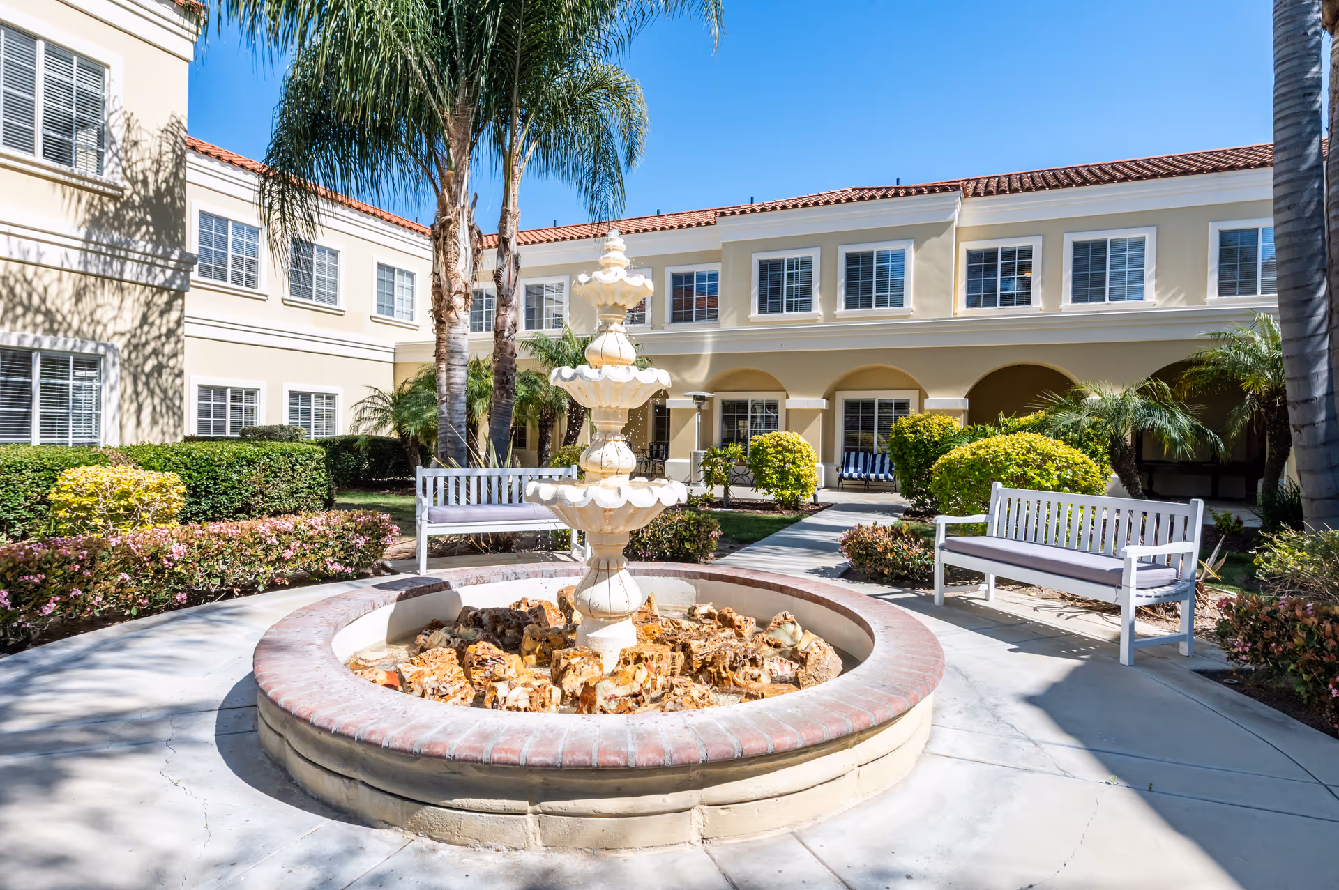Outdoor courtyard area at Capistrano Senior Living featuring a multi-tiered white fountain surrounded by a circular brick and stone base. The courtyard is landscaped with palm trees, bushes, and flowering plants. Two white benches with cushions are placed on either side of the fountain. The building surrounding the courtyard has beige walls, white window frames, and a red tile roof under a clear blue sky.