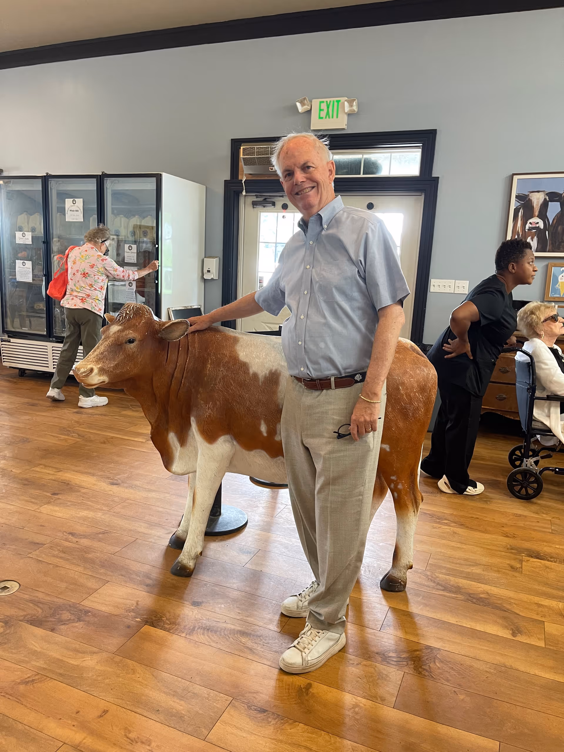 An elderly man smiling and standing indoors next to a life-sized model of a brown and white cow. In the background, a woman with a red backpack is looking at items in a glass refrigerator, and two other people are seated and standing near a wall with framed pictures. The room has wooden flooring and a door with an exit sign above it.