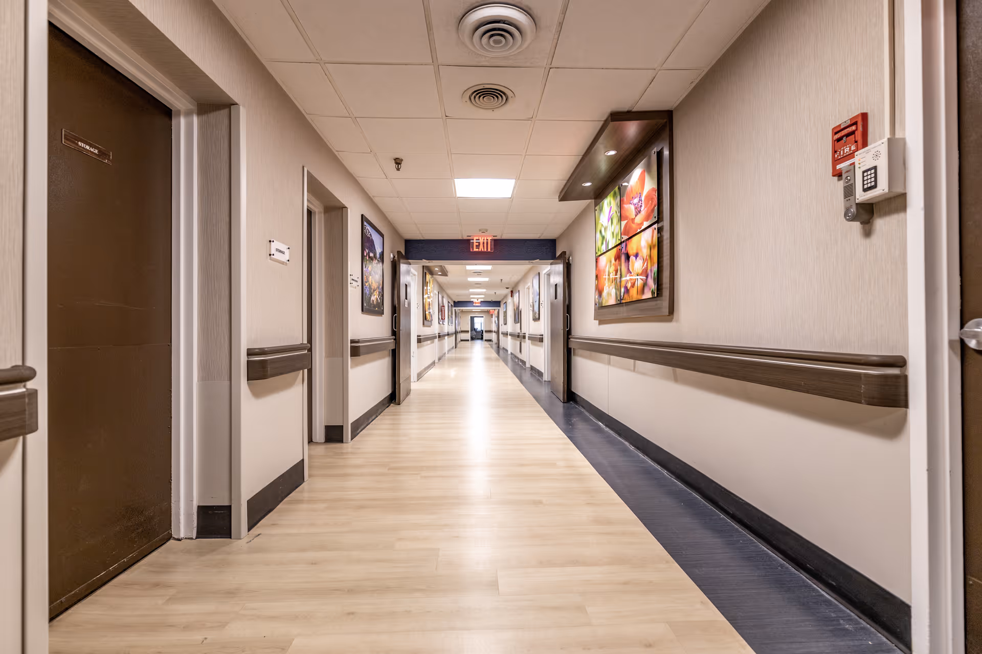A long, well-lit hallway in a senior living facility with light wood flooring, beige walls, and handrails on both sides. Several doors and framed pictures are visible along the walls. A fire alarm and keypad are mounted on the right wall near the foreground. The ceiling has recessed lighting and ventilation fixtures.