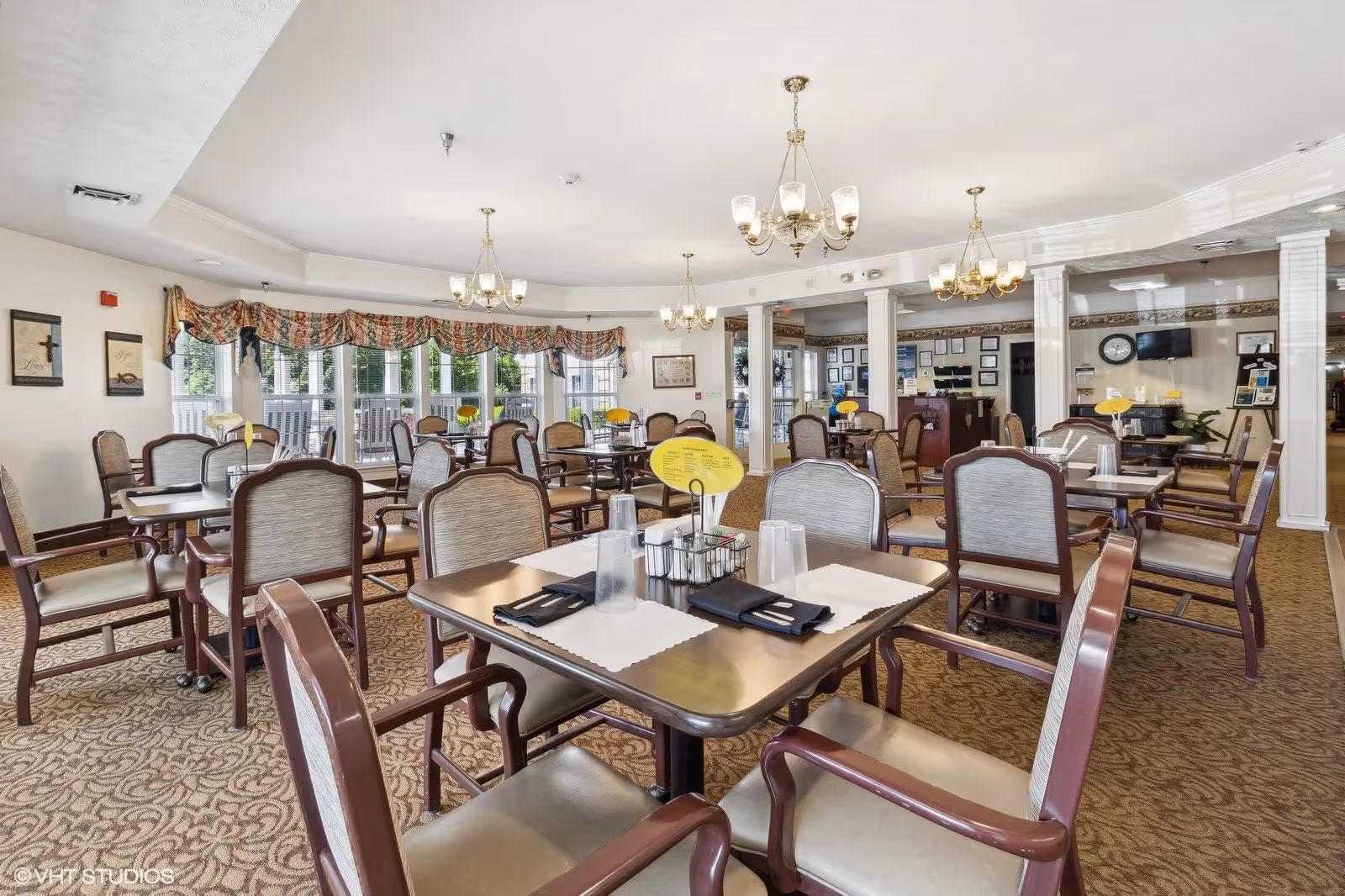 Bright, carpeted dining room with multiple set tables and chairs, chandeliers, and large windows in a senior living facility.