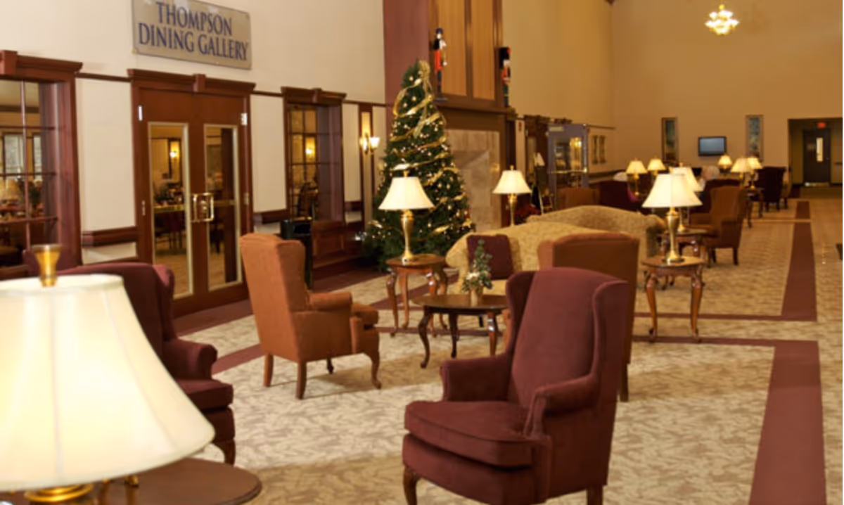 A spacious and warmly lit common area with multiple upholstered armchairs and small tables with lamps. A decorated Christmas tree stands near a fireplace, and double doors labeled 'Thompson Dining Gallery' are visible on the left side of the room.