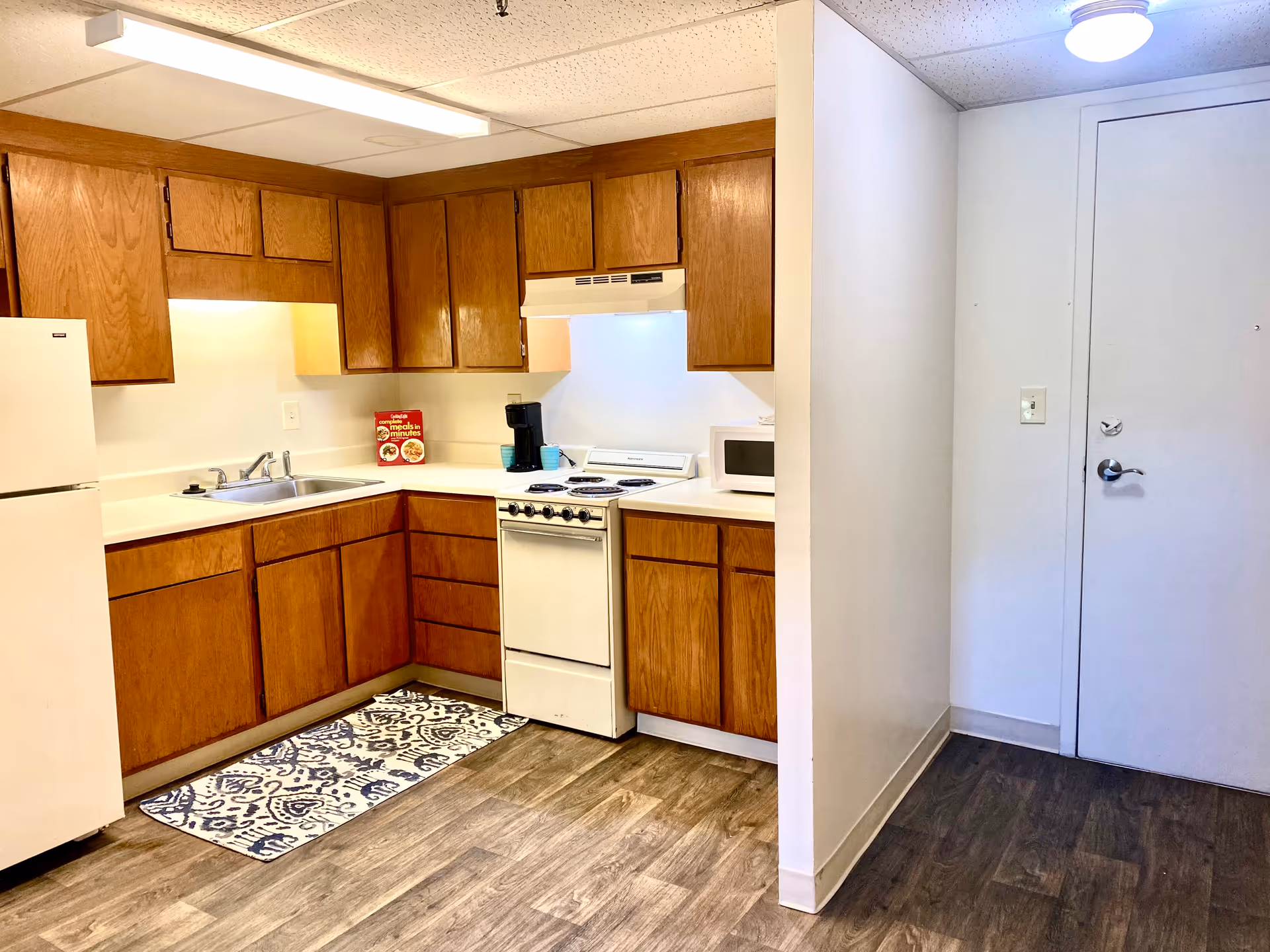 A small kitchen area with wooden cabinets, a white refrigerator, a white stove with an overhead vent, a microwave, a coffee maker, and a sink. The floor has a wood-like finish with a patterned rug in front of the sink. To the right is a white door and a small hallway area.