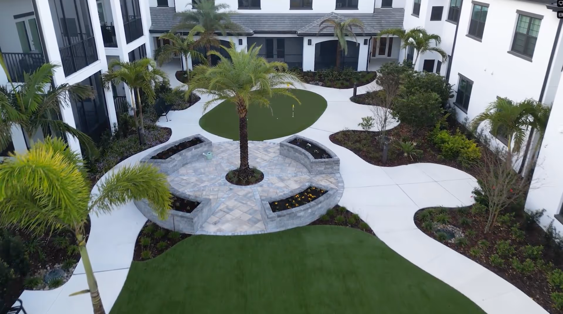 A landscaped outdoor courtyard area in a senior living facility featuring a circular stone seating area with a palm tree in the center, surrounded by green artificial turf and additional palm trees. The courtyard is enclosed by a white multi-story building with multiple windows and balconies.