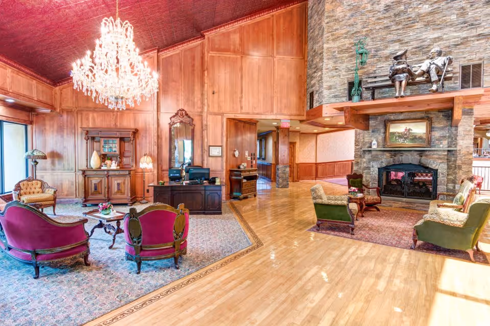 A spacious and elegant living room area with polished wooden floors and wood-paneled walls. The room features a large crystal chandelier hanging from a decorative ceiling, vintage-style furniture including green and purple upholstered chairs and sofas arranged around coffee tables, and a stone fireplace with a painting above it. There are statues of two people sitting on a bench on the mantel. The room is well-lit with natural light coming through large windows on the left side.