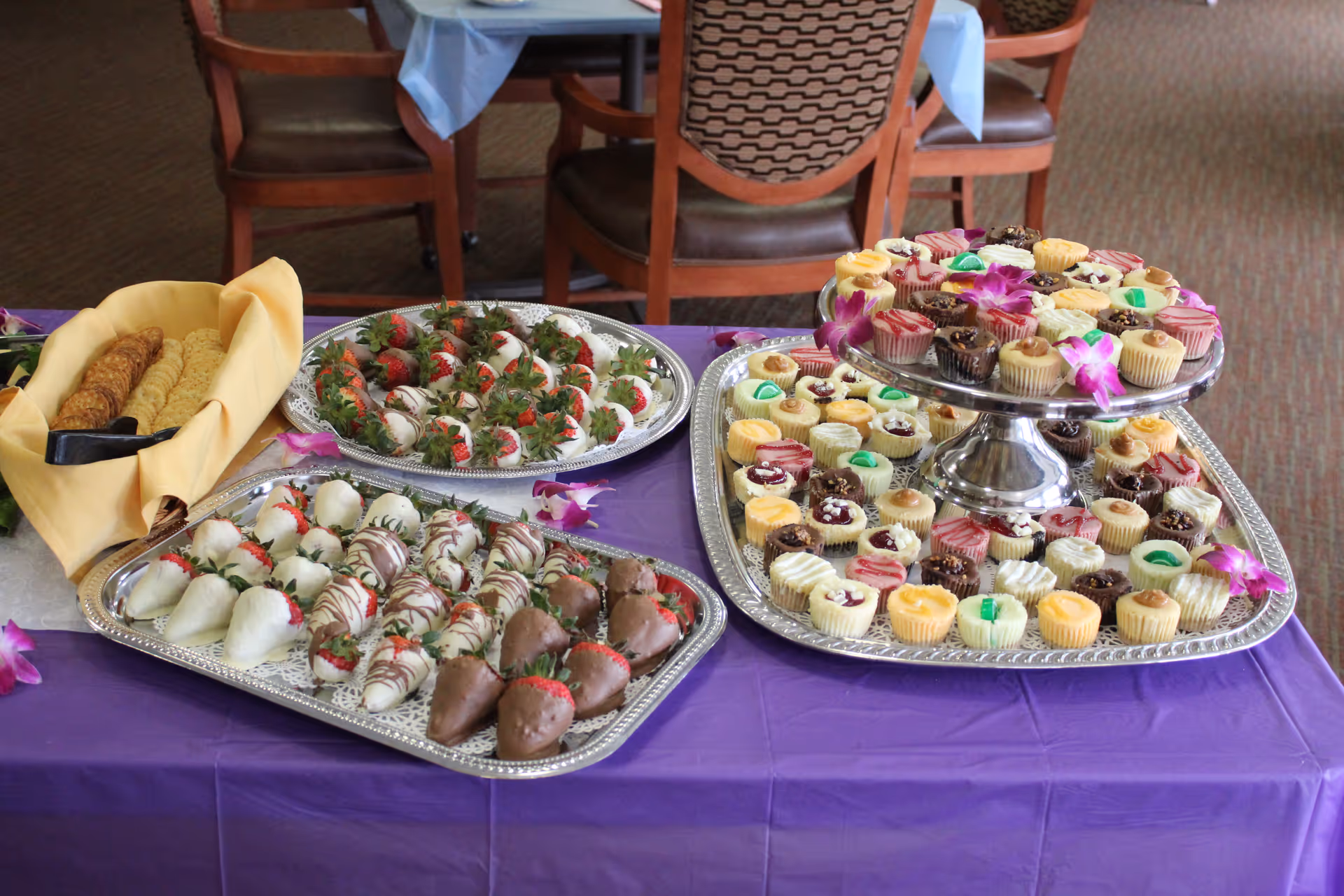 A table covered with a purple tablecloth displaying trays of assorted desserts including chocolate-covered strawberries, mini cheesecakes with various toppings, and a basket of crackers. The background shows wooden chairs and tables in a dining area.