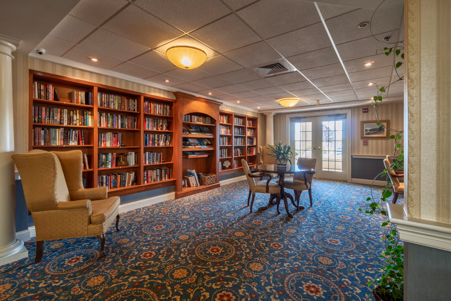 A cozy library or reading room with wooden bookshelves filled with books along the left wall, a patterned carpet, a round table with four upholstered chairs in the center, and a comfortable armchair to the left. There are large windows and glass doors at the far end letting in natural light, with some plants adding greenery to the space.