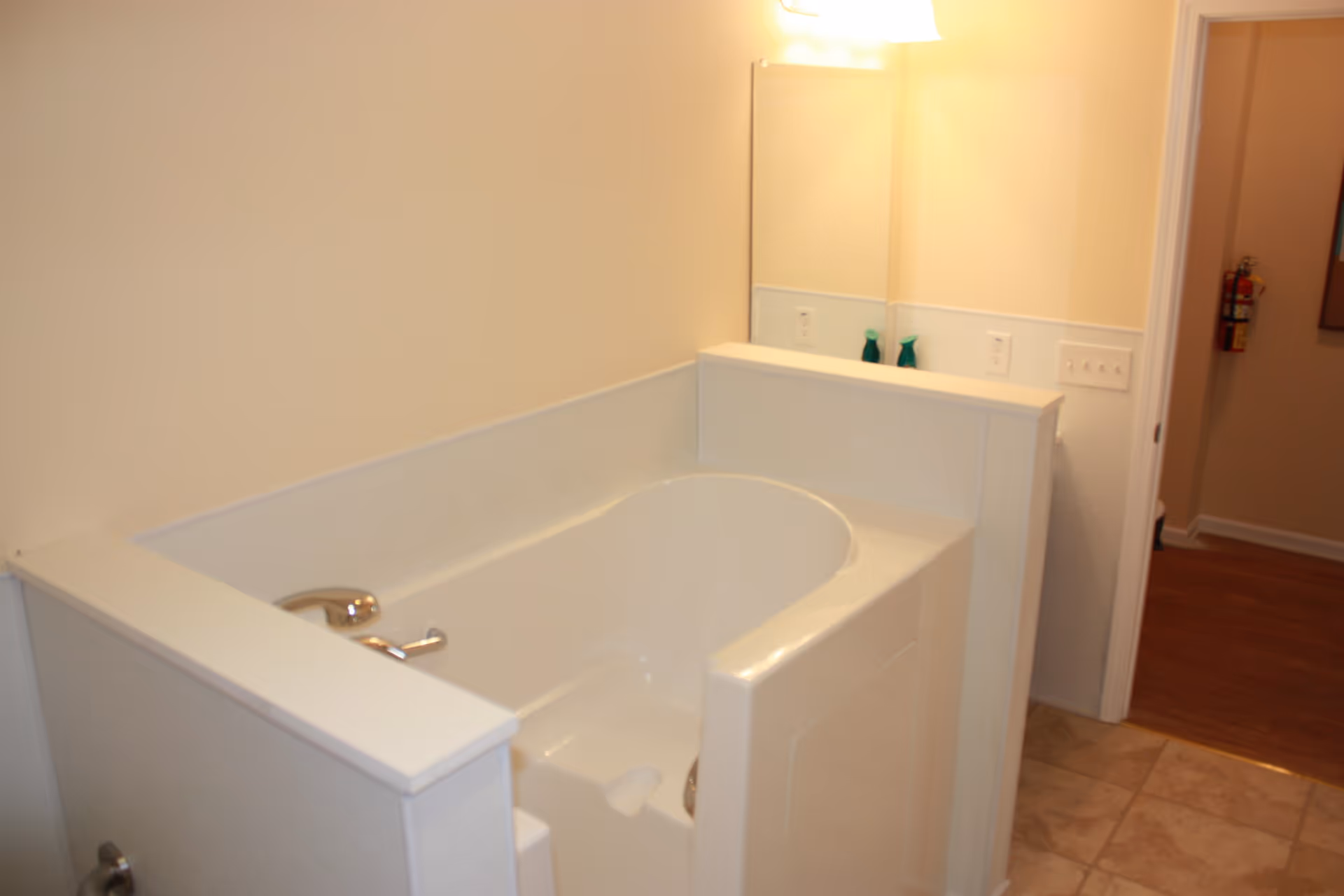 A walk-in bathtub in a bathroom with beige walls and tiled floor. The tub has a built-in seat and a faucet on the left side. A large mirror and a light fixture are visible above the tub. A fire extinguisher is mounted on the wall in the adjacent hallway.