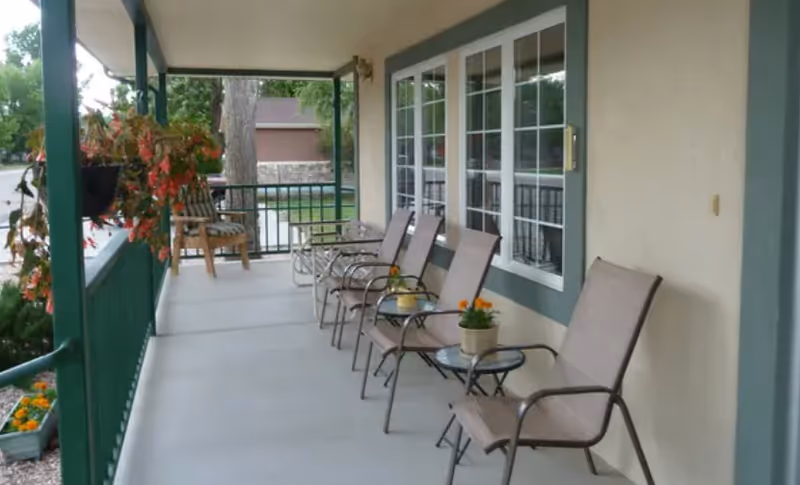 Covered outdoor porch area with several chairs and small tables, some with potted flowers, green railing, and a hanging plant. The porch overlooks a garden and a tree is visible in the background.