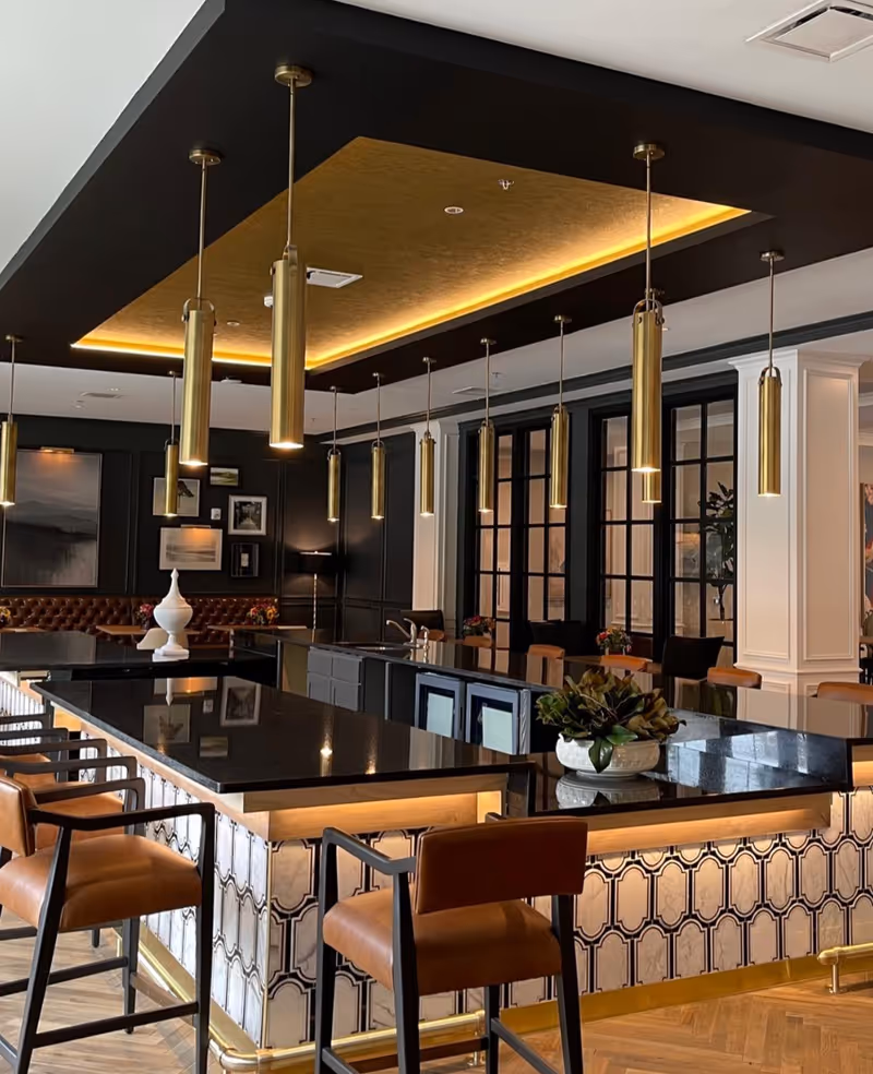 A modern and elegant interior space featuring a large black granite countertop island with patterned tile sides, surrounded by brown leather bar stools with black frames. Above the island, multiple cylindrical gold pendant lights hang from the ceiling, which has a recessed section with warm yellow lighting. The background includes dark paneled walls adorned with framed artwork and a tufted brown leather bench, as well as large black-framed glass doors and white columns.