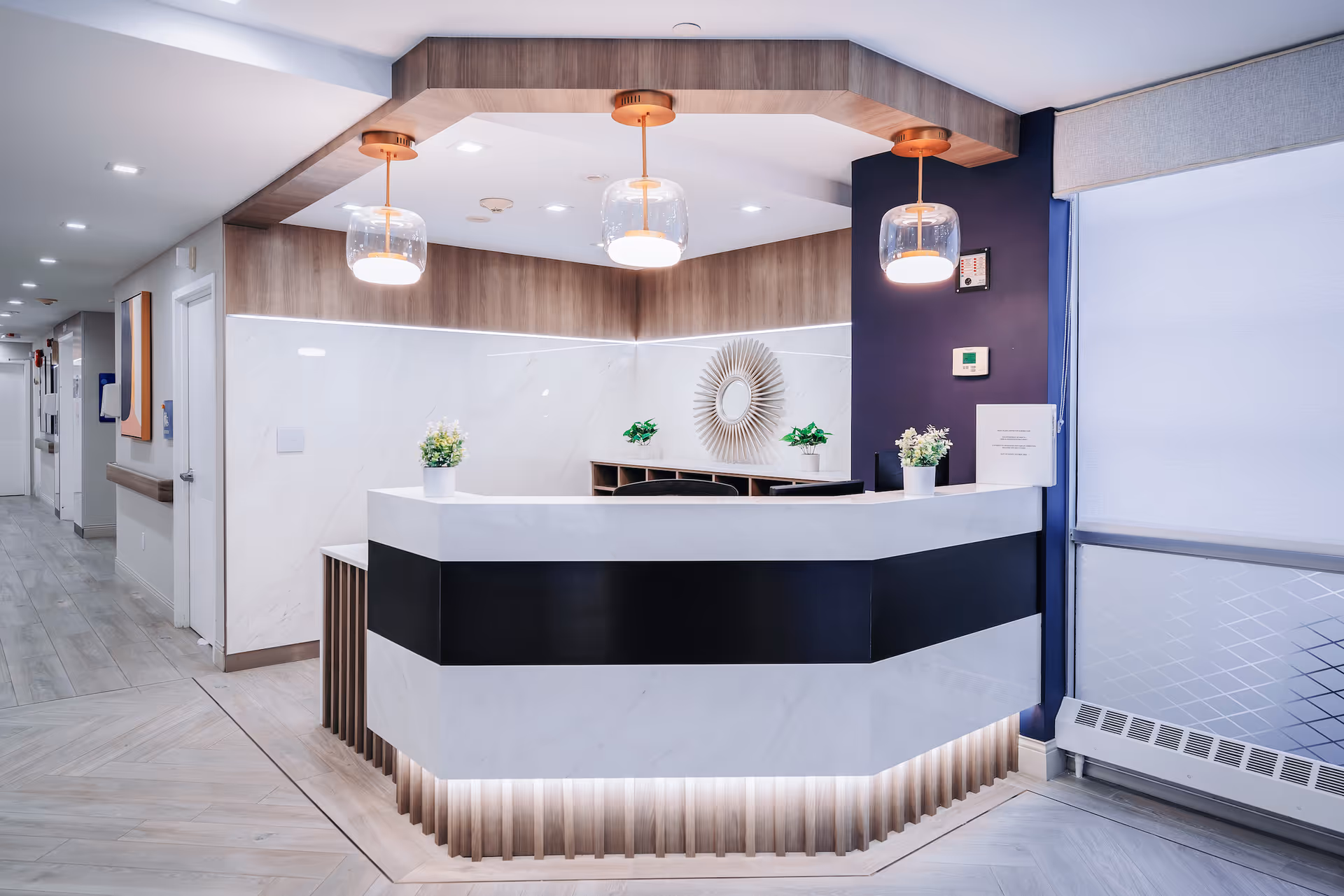 A modern reception desk area in a nursing care facility with a white and black counter, three pendant lights hanging from a wooden ceiling feature, potted plants on the counter, and a decorative wall mirror behind the desk. The floor is light wood, and there is a hallway with doors and artwork visible to the left.