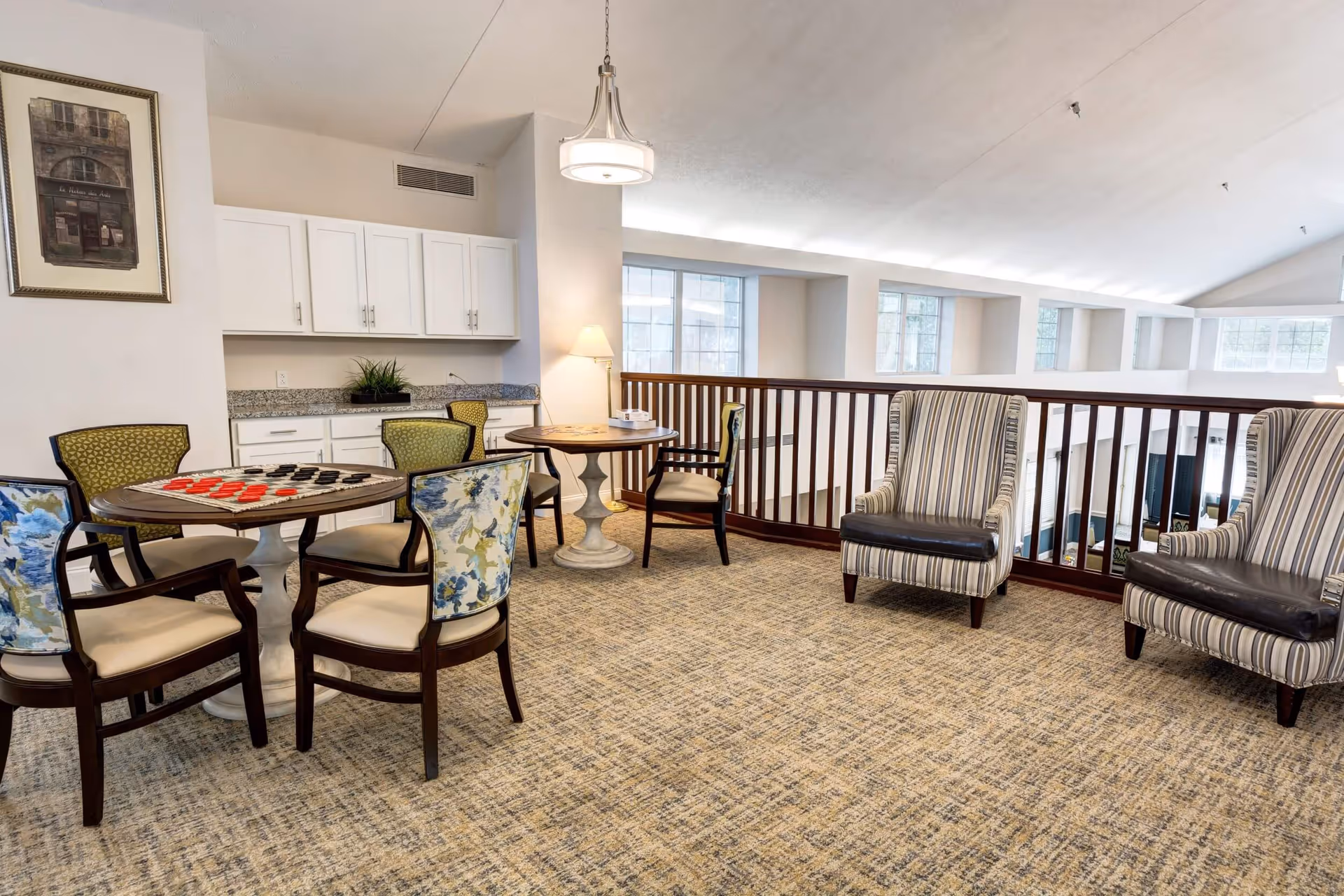 A cozy common area with a carpeted floor featuring two round tables surrounded by chairs, one table set up with a checkers game. The room has white cabinetry with a granite countertop, a framed picture on the wall, a hanging light fixture, and two striped armchairs near a wooden railing overlooking a lower level with large windows letting in natural light.