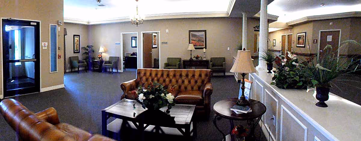 Interior view of a nursing and rehabilitation facility lobby with brown leather sofas, a coffee table with a flower arrangement, side tables with lamps, potted plants, and chairs along the walls. There is a glass door entrance on the left and several doorways leading to other rooms.